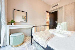 A bedroom with a bed, a desk, a chair, a small stool, a potted plant, and a door leading to another room. - Stylish 1 BR + Pool & World Class Gym (Dubai)