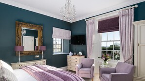 Ein Schlafzimmer mit einem Bett, zwei Sesseln, einem Schminktisch, einer Kommode, einem Spiegel, einer Chandelüster und einem Fenster mit Vorhängen.