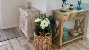 Una habitación con un aparador de madera, una cesta de mimbre con flores, una consola de madera con un jarrón y dos espejos colgados en la pared.