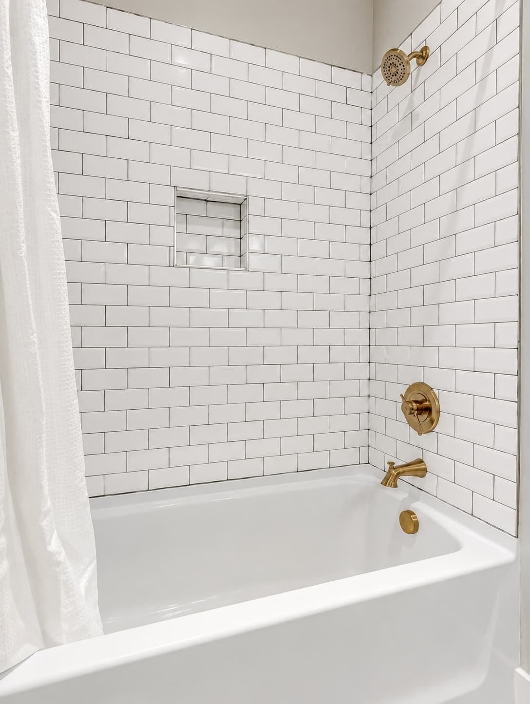 A bathroom with white subway tile walls, a white bathtub, and gold fixtures.