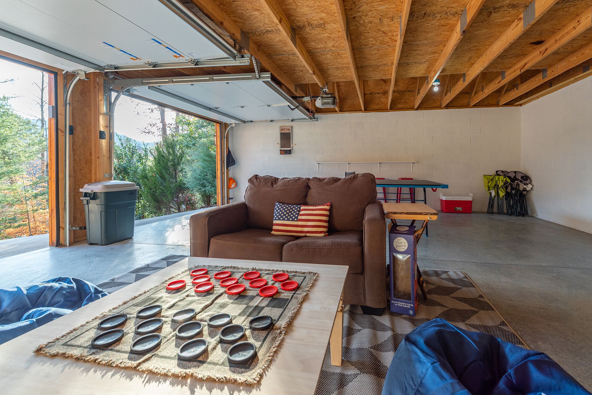 Eine geräumige Garage mit Holzbalkendecke, einem braunen Ledersofa mit Kissen mit US-Flagge, einem Tisch mit einem Gesellschaftsspiel und Blick auf Bäume durch die offene Garagentür.