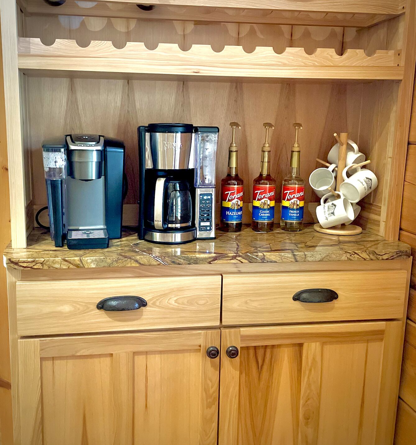 Una cocina de madera con una cafetera, dos botellas de jarabe Torani y un soporte para tazas.