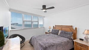 Un dormitorio con cama, ventilador de techo, mesita de noche, lámpara, silla y una pequeña planta.