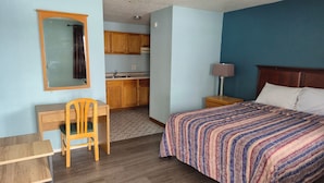 A bedroom with a bed, a desk, a chair, a nightstand, a lamp, and a kitchenette in the background. - Napp Inn Motel (Rock Falls)