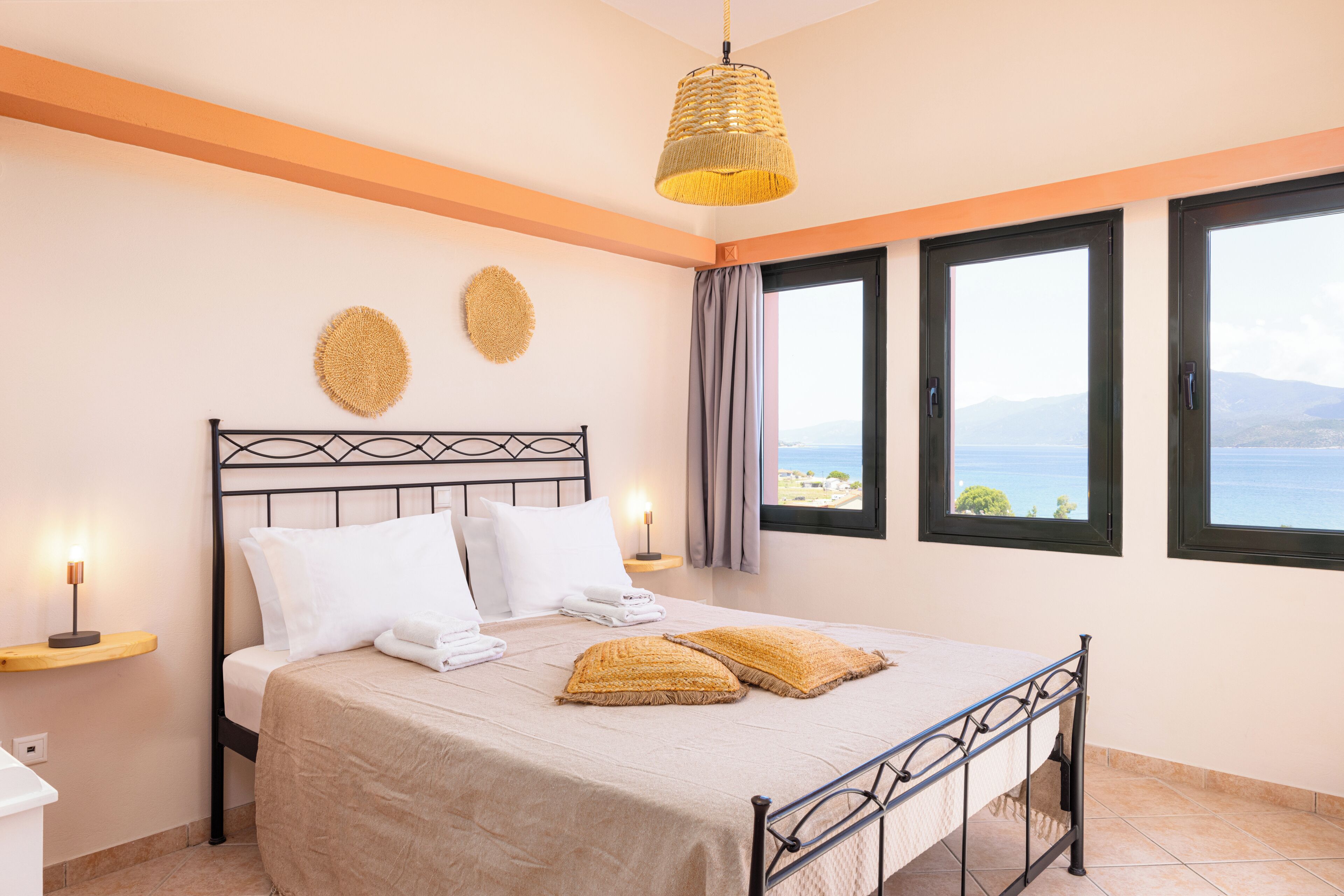 A neatly made bed with a beige bedspread, two pillows, and two decorative pillows. There are two bedside tables with lamps, a window with a view of the sea, and a woven pendant light above the bed.