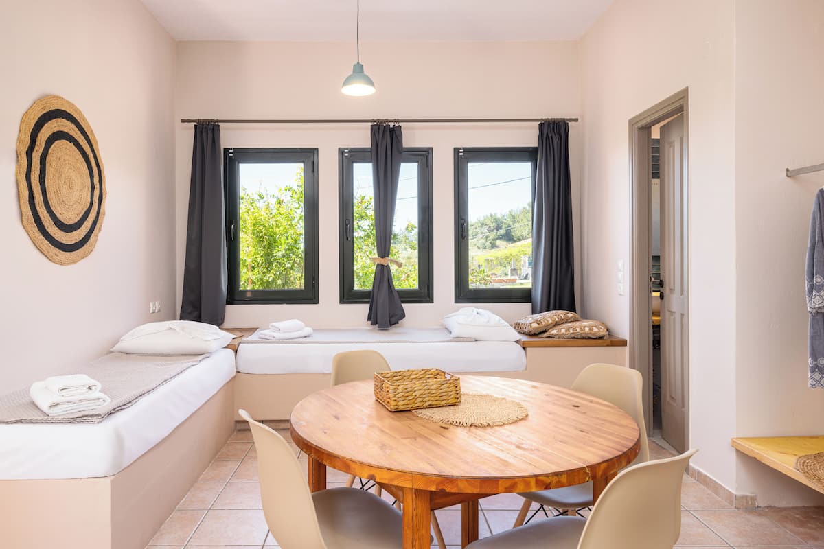 A room with a bed, a dining table, chairs, a window with curtains, a towel rack, and a basket on the table.