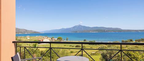 A balcony with a table and chairs, overlooking a coastal landscape with mountains and a clear sky.