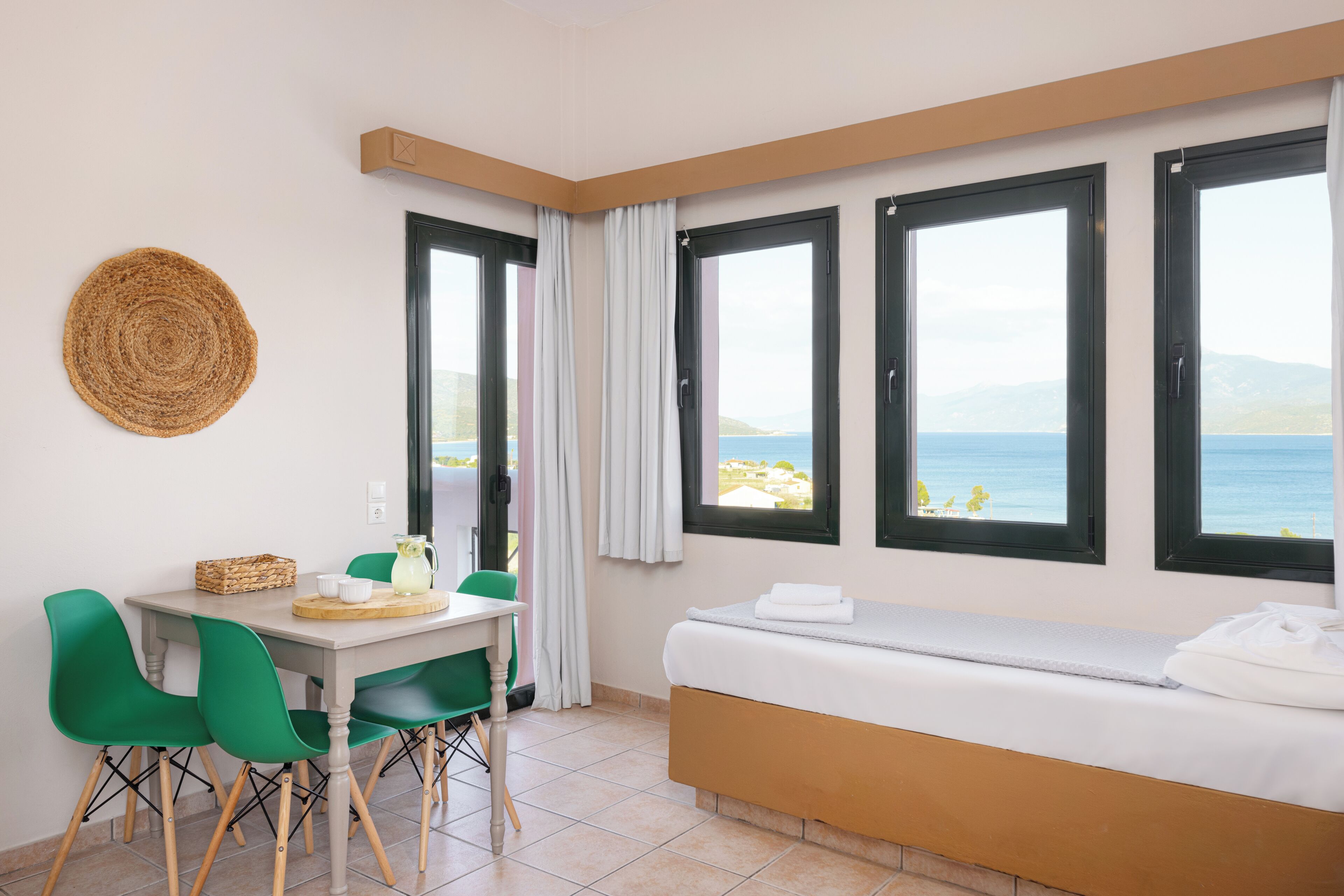 A room with a bed, a dining table with chairs, and large windows with a view of the sea.
