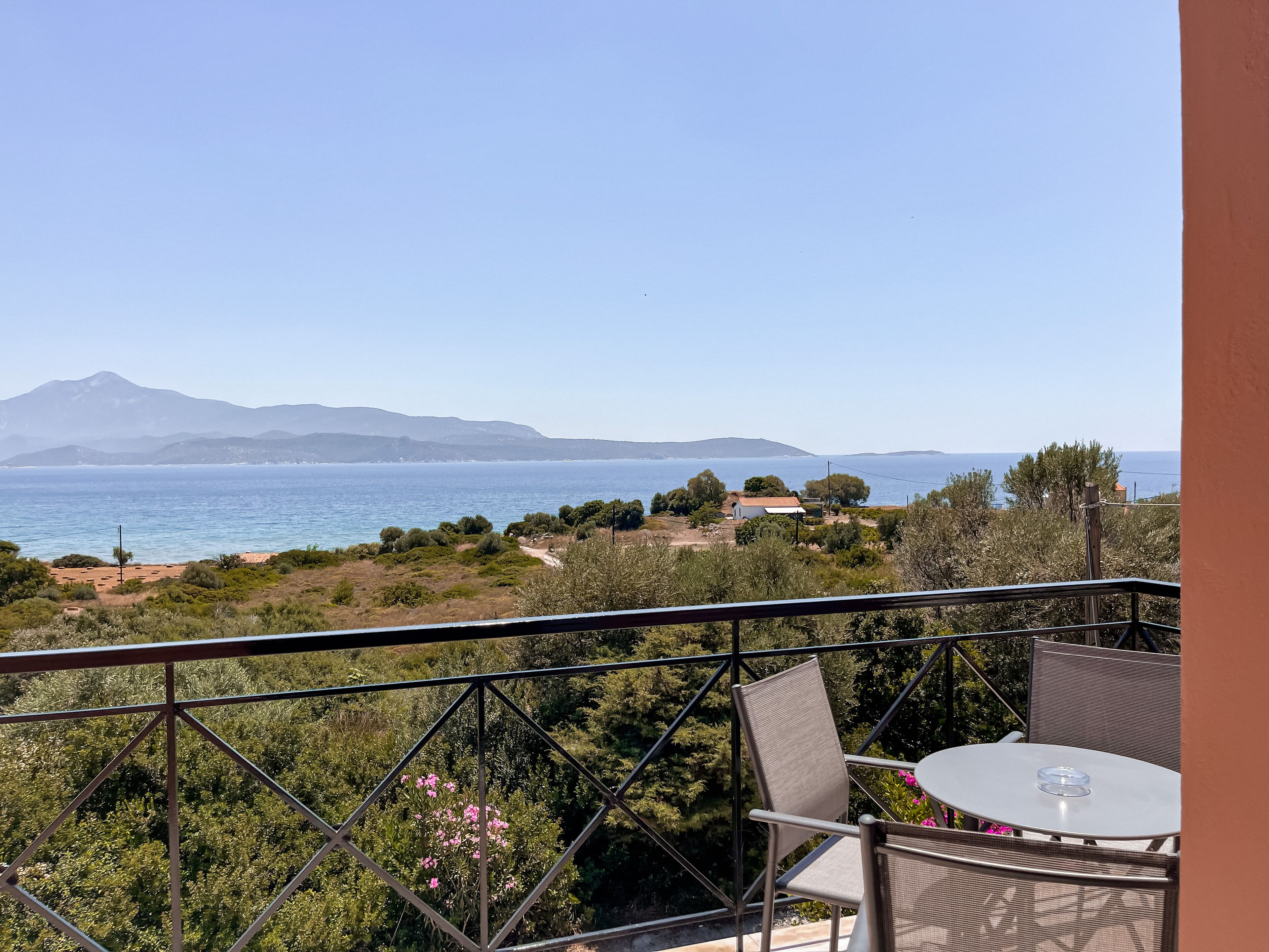 Un balcón con mesa y sillas, con vistas a un paisaje costero y una vista despejada del mar y montañas distantes.