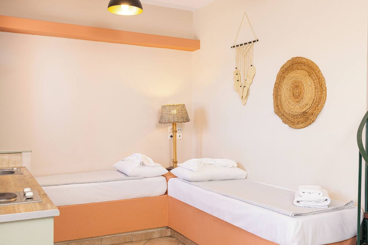 A small, cozy room with two single beds, a bedside table, a lamp, and a wall-mounted macrame decoration.