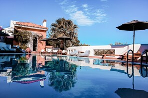 A clear swimming pool with lounge chairs and umbrellas, surrounded by buildings with red-tiled roofs and palm trees.
