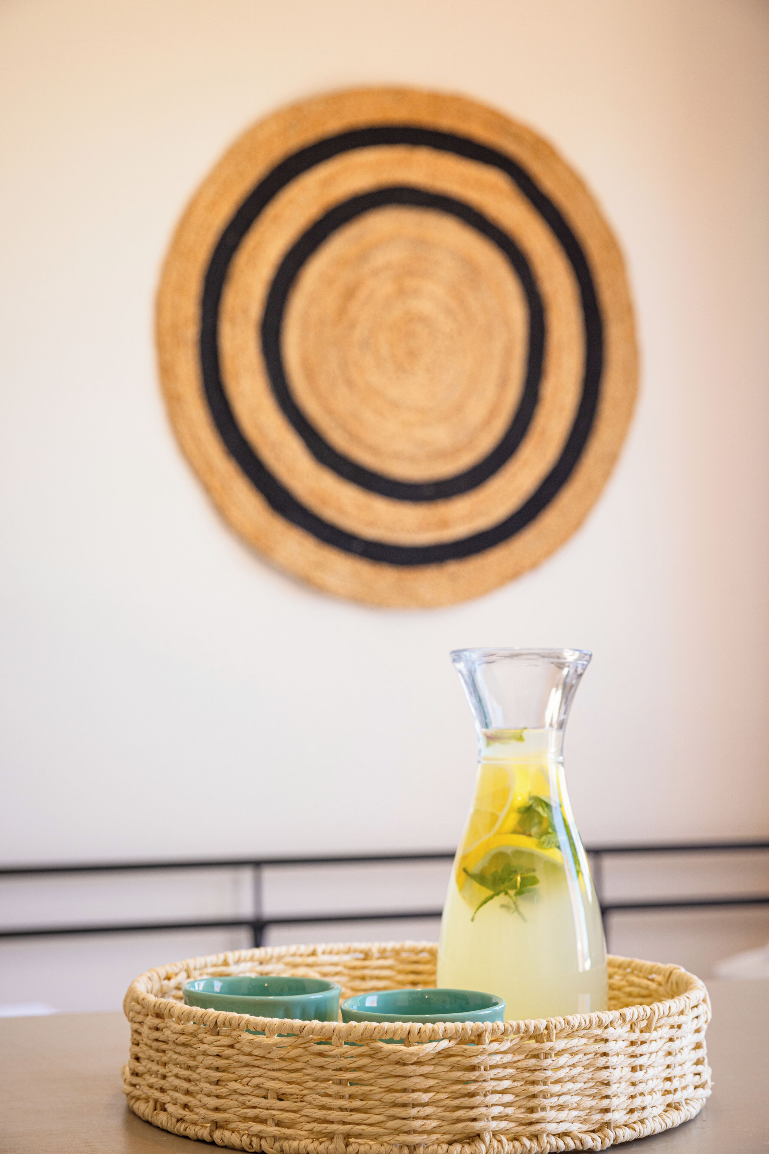 Een gevlochten mand met een glazen karaf gevuld met een geel drankje, vergezeld van twee blauwgroene bekers, geplaatst op een tafel tegen een muur met een ronde gevlochten mat.