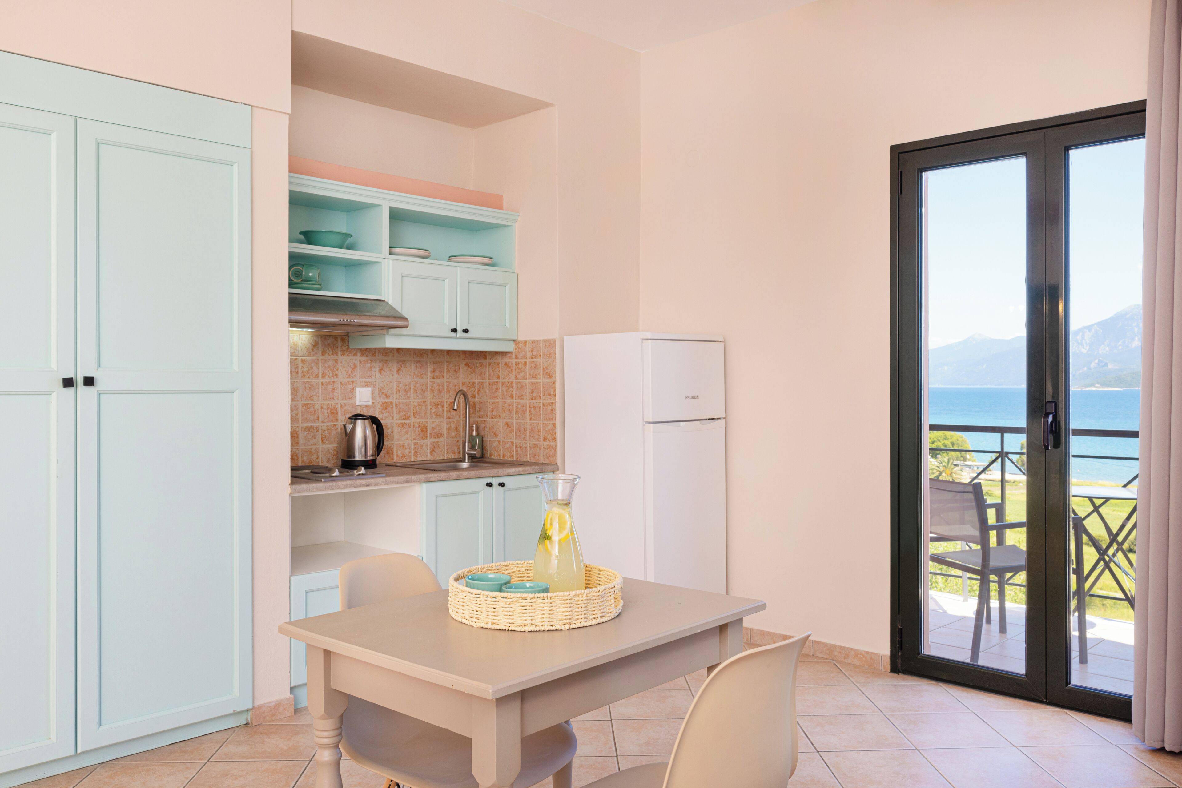 A kitchen with light blue cabinets, a white refrigerator, and a dining table with chairs. There is a window with a view of the sea and mountains.