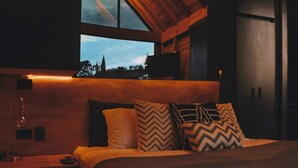 Une chambre confortable avec une grande fenêtre, des poutres en bois au plafond et un lit agrémenté d’oreillers à motifs.