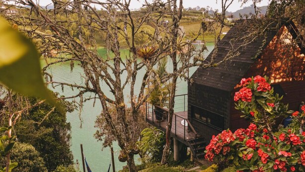 Une cabane en bois dotée d’une terrasse donnant sur un lac, entourée d’une végétation luxuriante et de fleurs rouges.