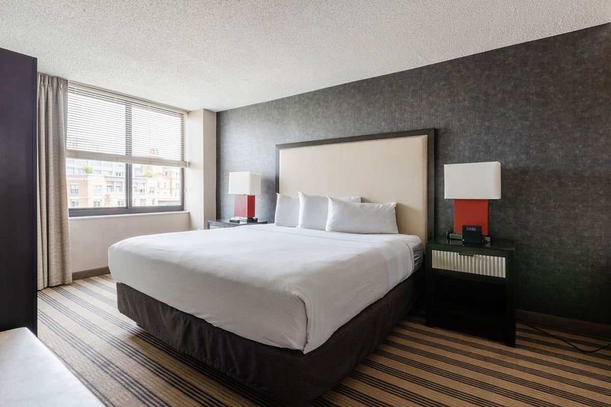 Quarto de hotel com uma cama grande, duas mesas de cabeceira com luminárias, uma janela com cortinas e um tapete listrado.
