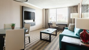 A modern hotel room with a sofa, a dining table with chairs, a TV mounted on the wall, and a large window with curtains. - 2500 Penn, a Placemakr Experience (Washington)
