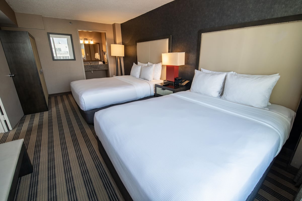 A hotel room with two beds, a desk, a chair, a wardrobe, a mirror, and a bathroom visible through an open door.