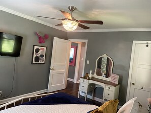 Une chambre à coucher comprenant un lit, une commode, un ventilateur de plafond, un téléviseur et une porte donnant sur une autre pièce.
