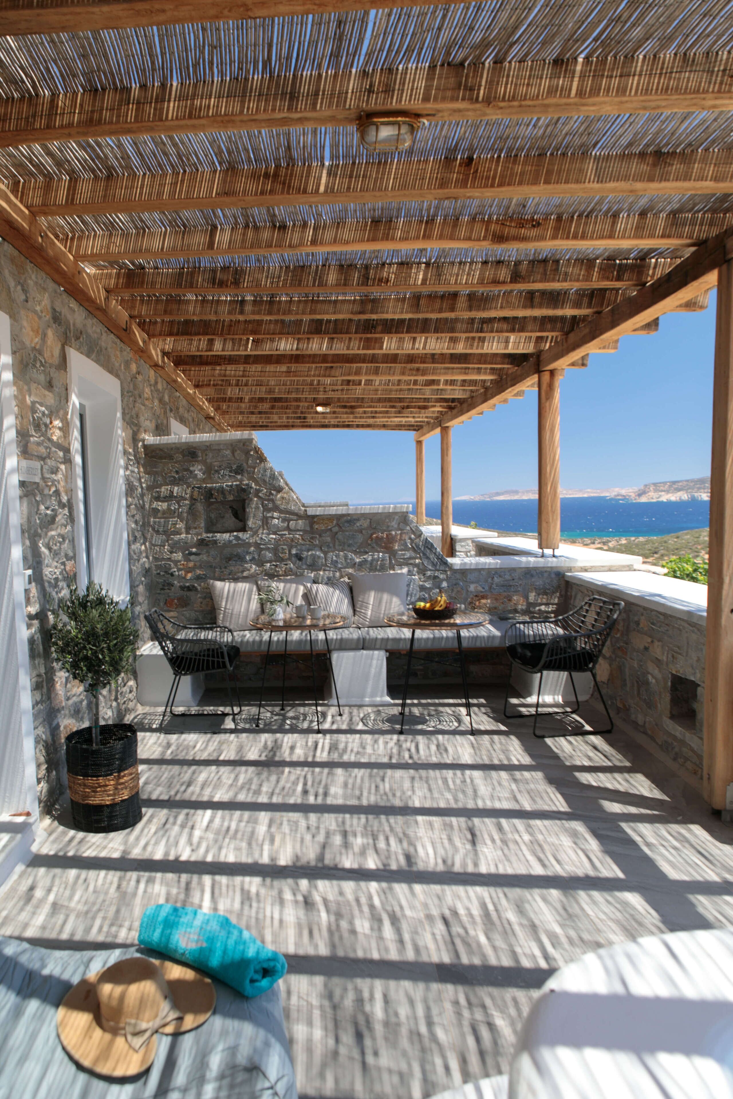 A stone-walled outdoor seating area with a view of the sea, featuring a pergola, cushions, and a table with fruit.