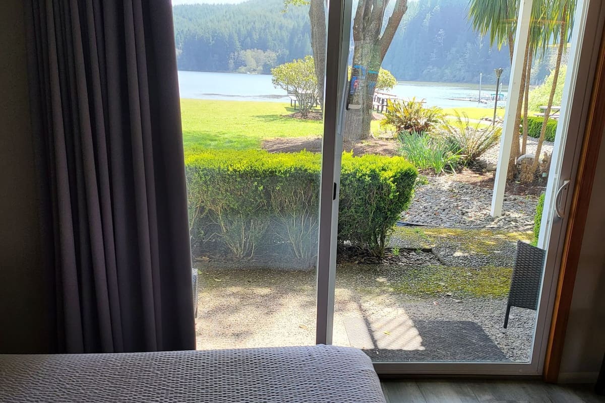 Ein Zimmer mit einer großen Schiebetür, die auf einen Balkon mit Blick auf einen See, viel Grün und ein fernes Bergmassiv führt.