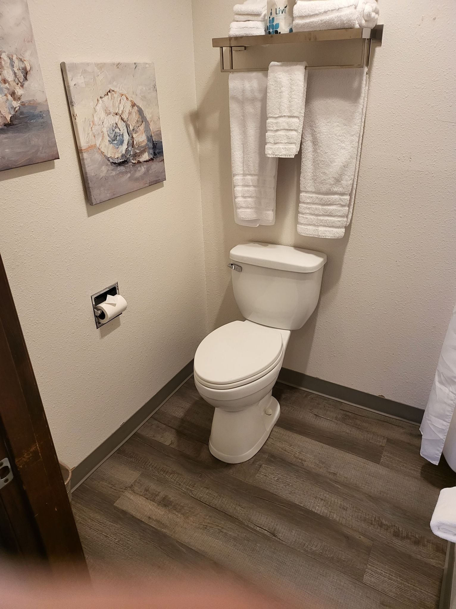A bathroom with a white toilet, a towel rack with folded towels, a painting on the wall, and a toilet paper holder.