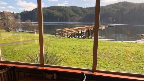 Un comedor con mesa y sillas de madera, ventanas amplias que dan a vistas de un lago, un muelle de madera y un área con césped.