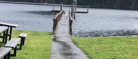 A concrete pathway leading to a wooden dock over a body of water, with picnic tables on the left and a grassy area on the right.