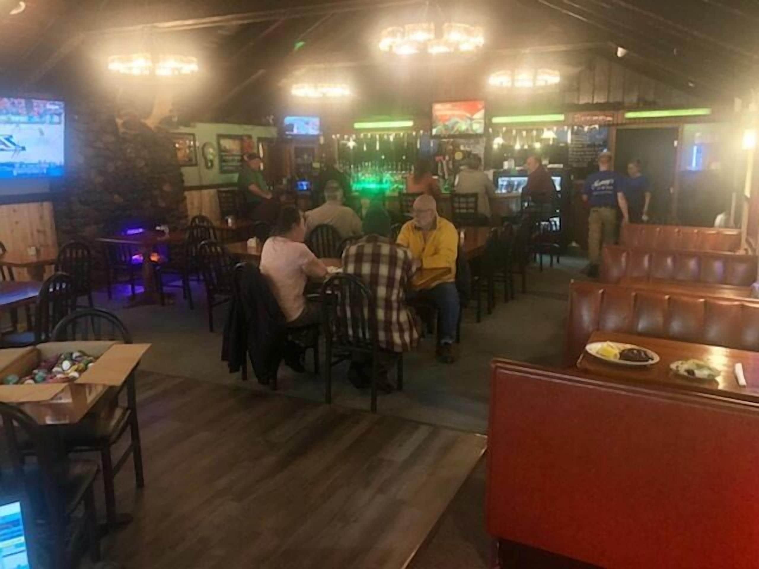 Interior de un restaurante con mesas y sillas de madera, una zona de bar con televisores y una pared de piedra.
