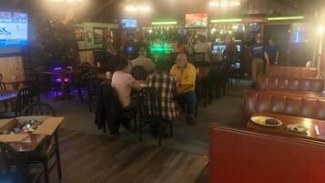 Interior de un restaurante con mesas y sillas de madera, una zona de bar con televisores y una pared de piedra.