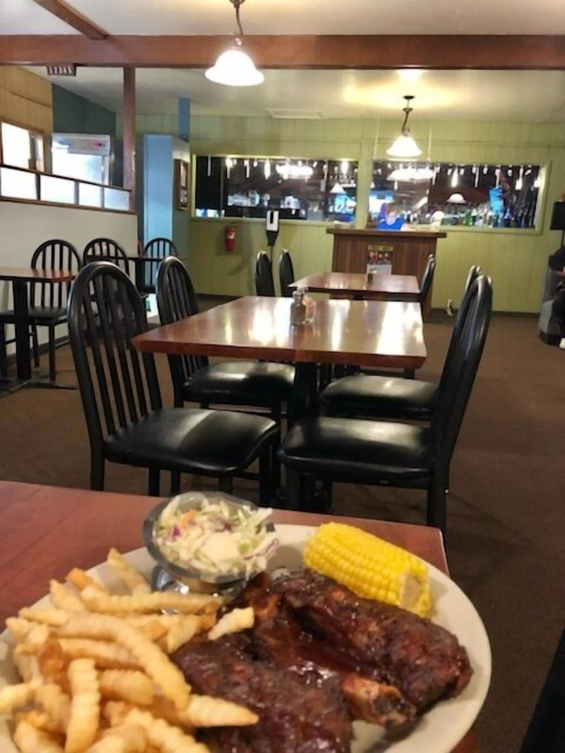 Interior de restaurante con mesas y sillas, una barra y un plato de comida que incluye costillas, maíz y papas fritas.
