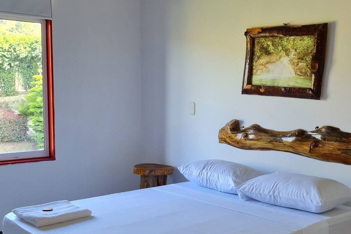 A bedroom with a large bed, a wooden headboard, a small bedside table, and a window with a view of greenery.