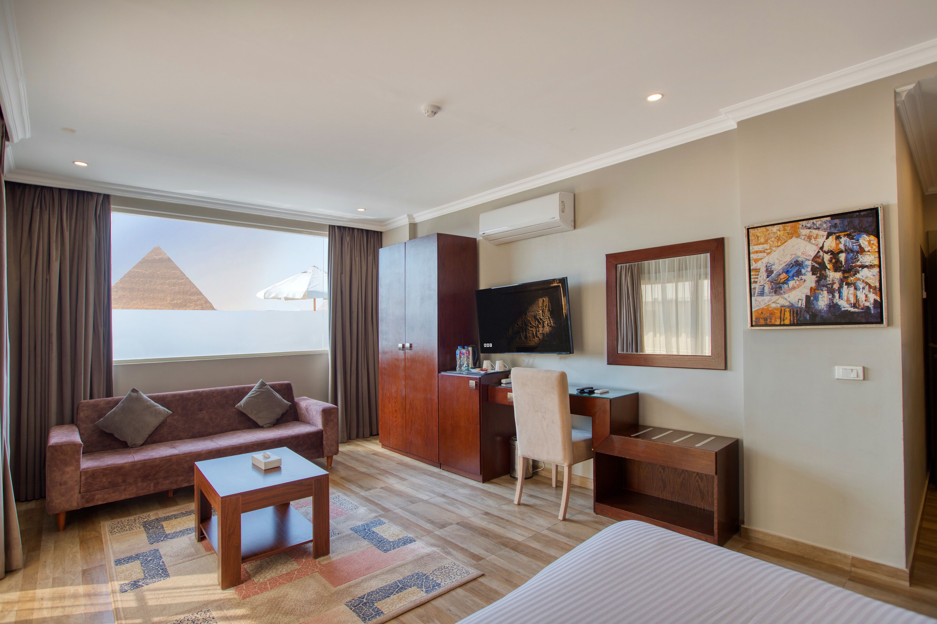 A hotel room with a sofa, a desk, a TV, a painting, and a view of the pyramid.