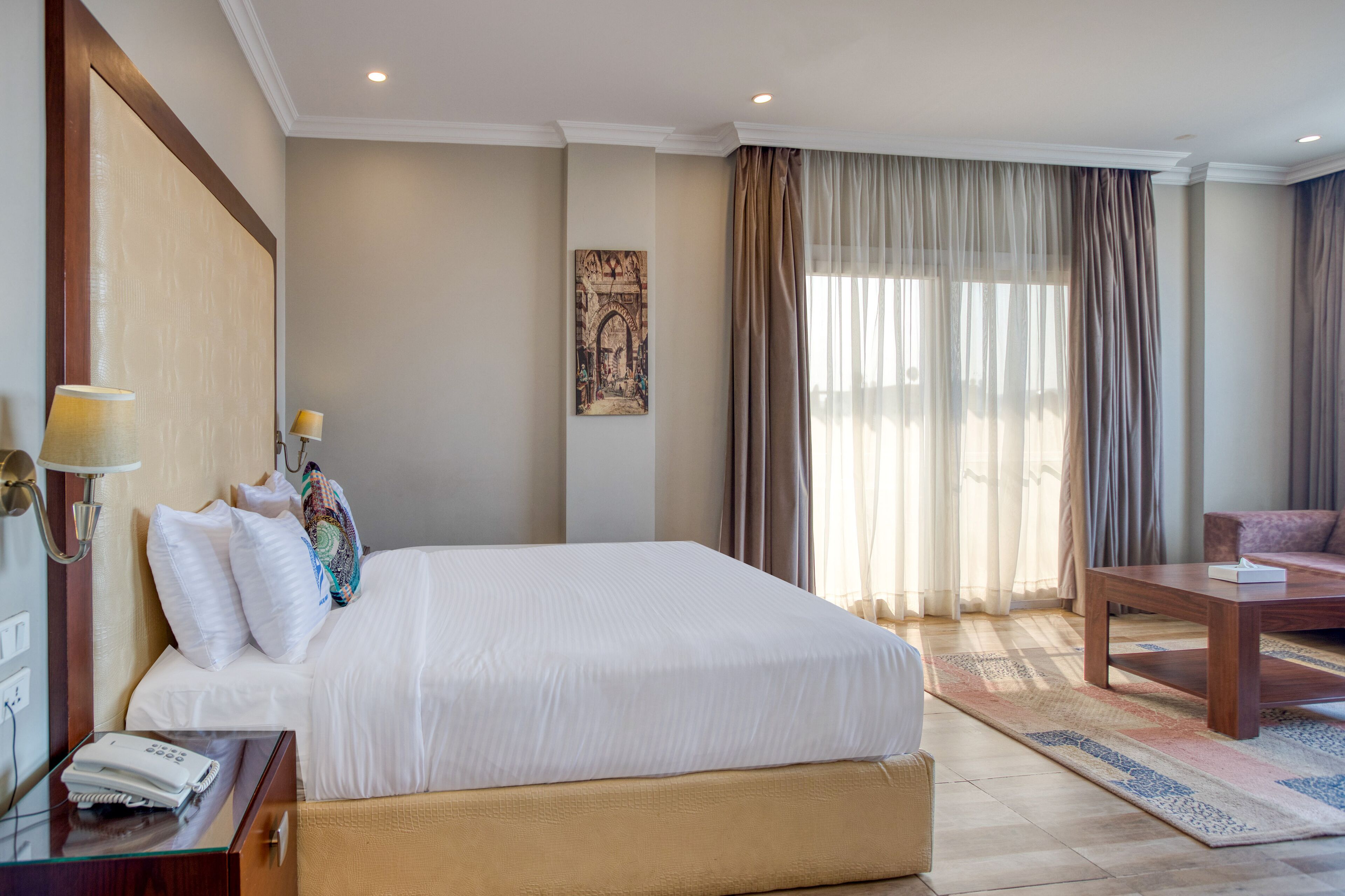Ein Hotelzimmer mit einem großen Bett, einem Nachttisch aus Holz, einem Schreibtisch, einem Sessel und einem großen Fenster mit Vorhängen.