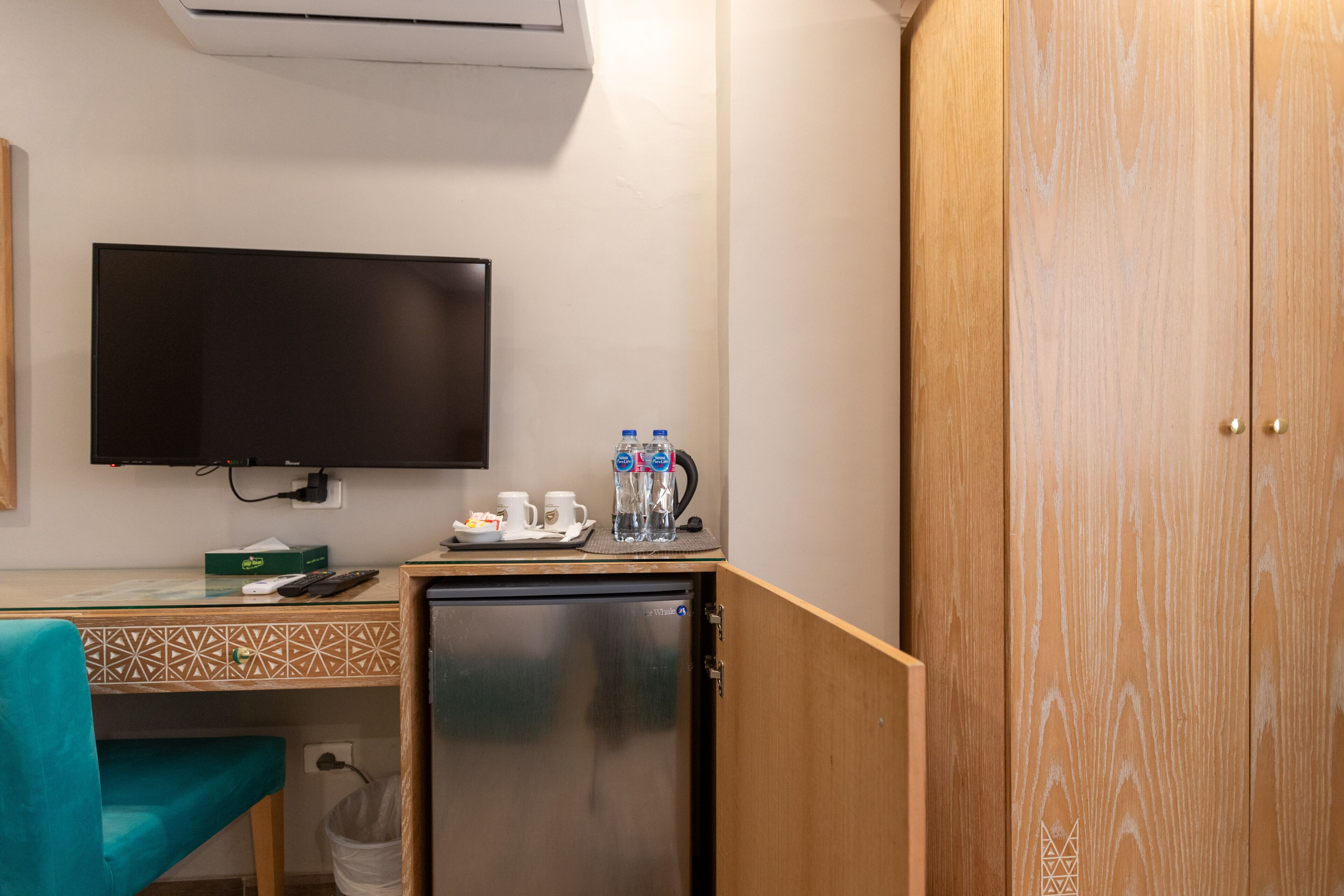 A compact room with a flat-screen TV, a small refrigerator, a wooden wardrobe, and a kitchenette area with a coffee maker and water bottles.