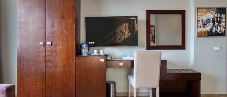 A room with a wooden wardrobe, a desk with a chair, a television, and a framed artwork on the wall.