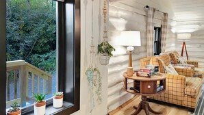Een gezellig interieur van een blokhut met een houten vloer, een ruitenpatroonbank, een kleintje tafeltje met een lamp en een raam met potplanten.