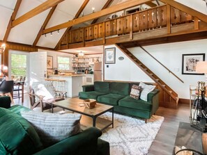 A spacious living area with a green sofa set, a wooden staircase, and a kitchen in the background.