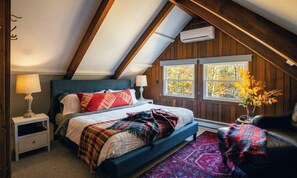 Una camera da letto con soffitto spiovente, un letto con numerosi cuscini, un comodino, una sedia e una finestra con foglie autunnali.