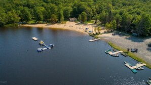 Vue aérienne d’une propriété au bord du lac, avec une plage de sable, des bateaux amarrés et un petit chalet.