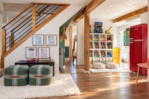 A spacious room with a wooden staircase, a bookshelf filled with books and toys, a red refrigerator, and a green sofa with striped cushions.