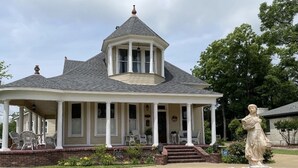 Een huis met twee verdiepingen, een veranda rondom, een torentje en een beeld in de voortuin.