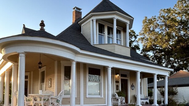 Een tweelaags huis met een veranda, een schoorsteen en een torentje op de tweede verdieping.