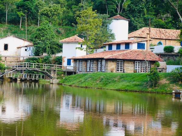 A traditional stone house with a tiled roof, a small bridge over a calm river, and lush greenery in the background.