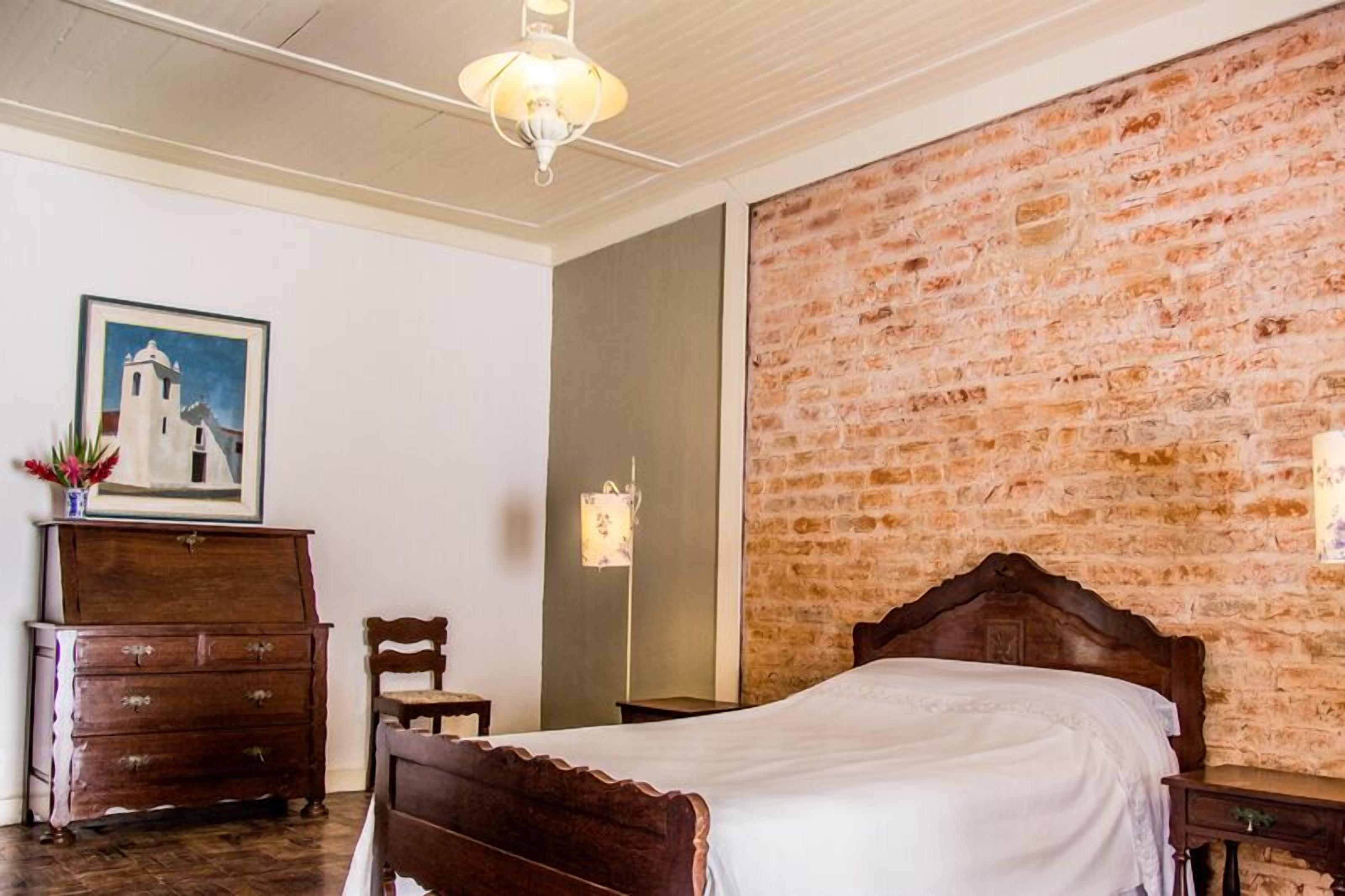 Ein Schlafzimmer mit einem hölzernen Bett, einem Schminktisch, einem Sessel, einem Gemälde und einer Ziegelwand.