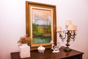 A framed landscape painting, a white vase with dried flowers, a small ornate container, and a candelabra with multiple candles on a wooden table against a white wall.