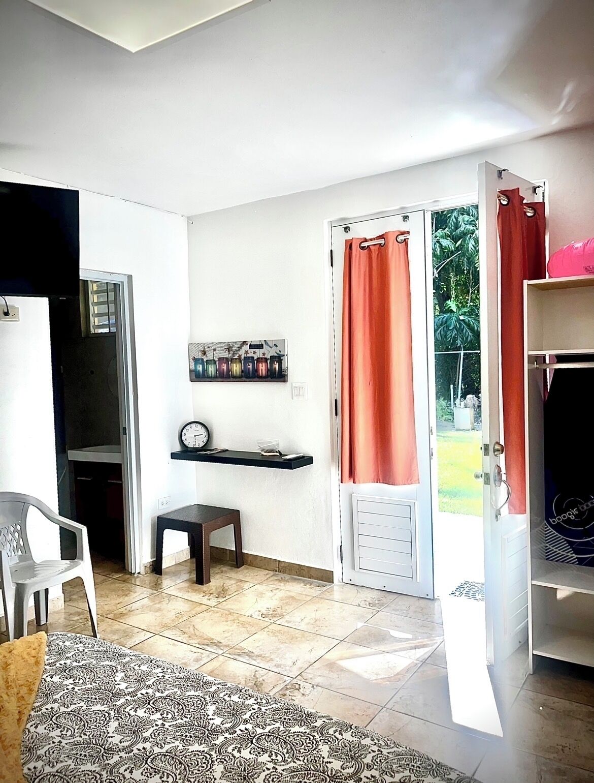 Un espace de vie compact comprenant un coin repas, une petite table, une chaise, une horloge, une étagère avec des livres et une porte coulissante donnant sur un jardin.