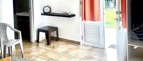 Un espace de vie compact comprenant un coin repas, une petite table, une chaise, une horloge, une étagère avec des livres et une porte coulissante donnant sur un jardin.