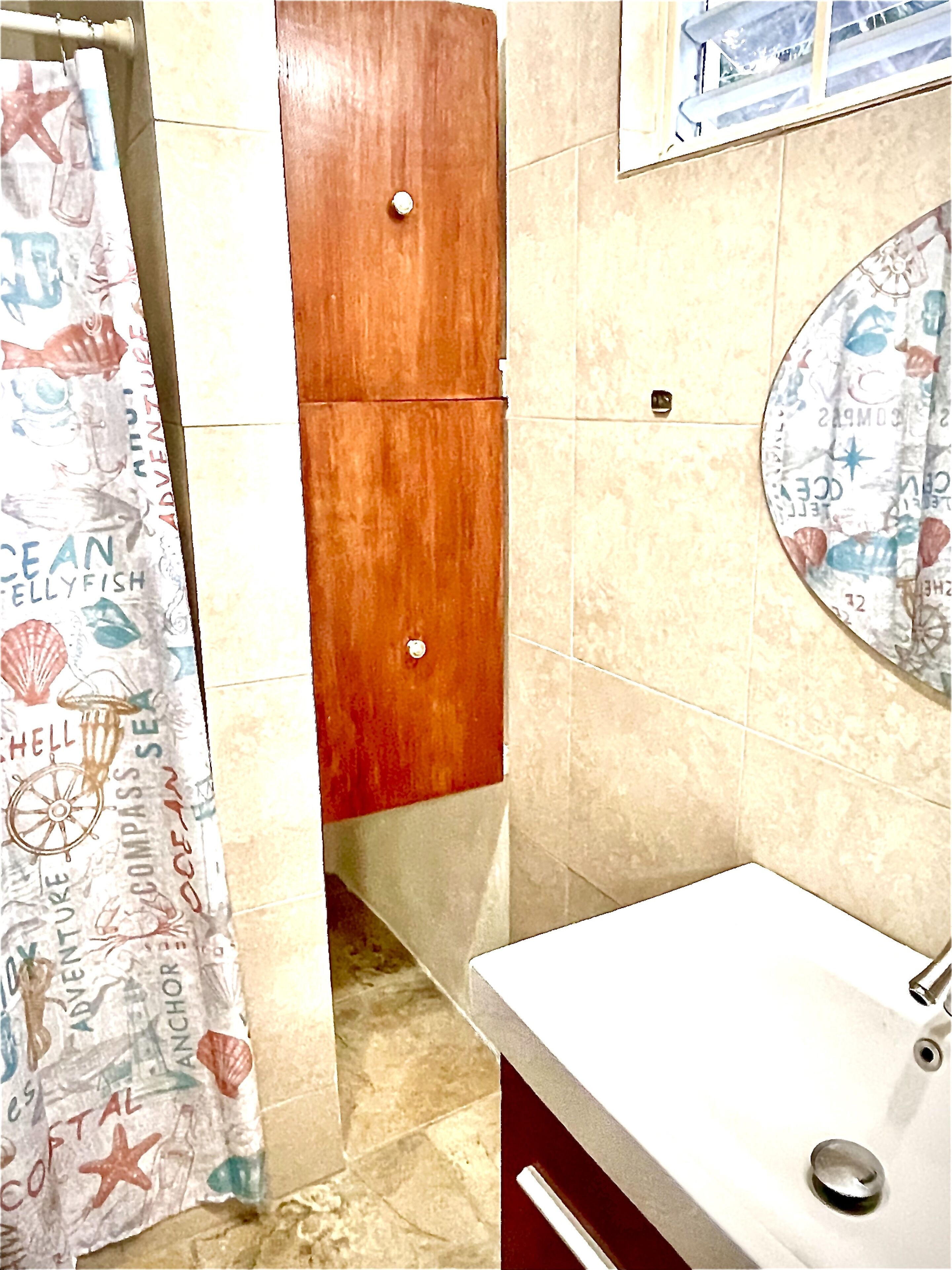 A bathroom with a wooden door, a tiled floor, a white sink, and a shower curtain with a nautical theme.
