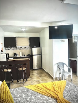 A compact kitchen with a refrigerator, stove, and bar stools. A flat-screen TV is mounted on the wall.